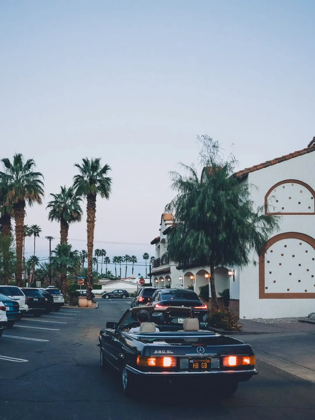 A vintage Mercedes 280SL convertible, top down, leaving a parking lot at dusk with a couple in the front seats. Spanish Colonial buildings with red tile roofs and palm trees behind. California plate "HB GB".