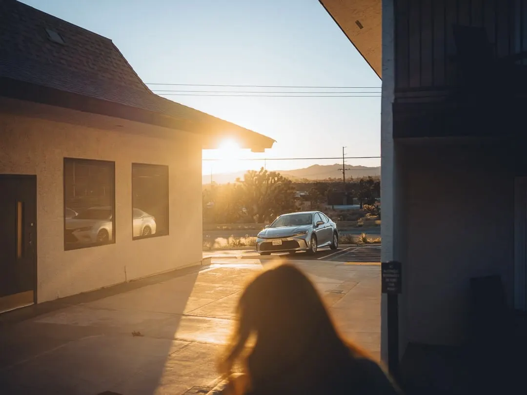 Low desert sun setting between two stucco buildings, a silver Toyota parked in the gap, Joshua trees and a ridge of low mountains in the middle distance. A person's silhouette in the foreground.