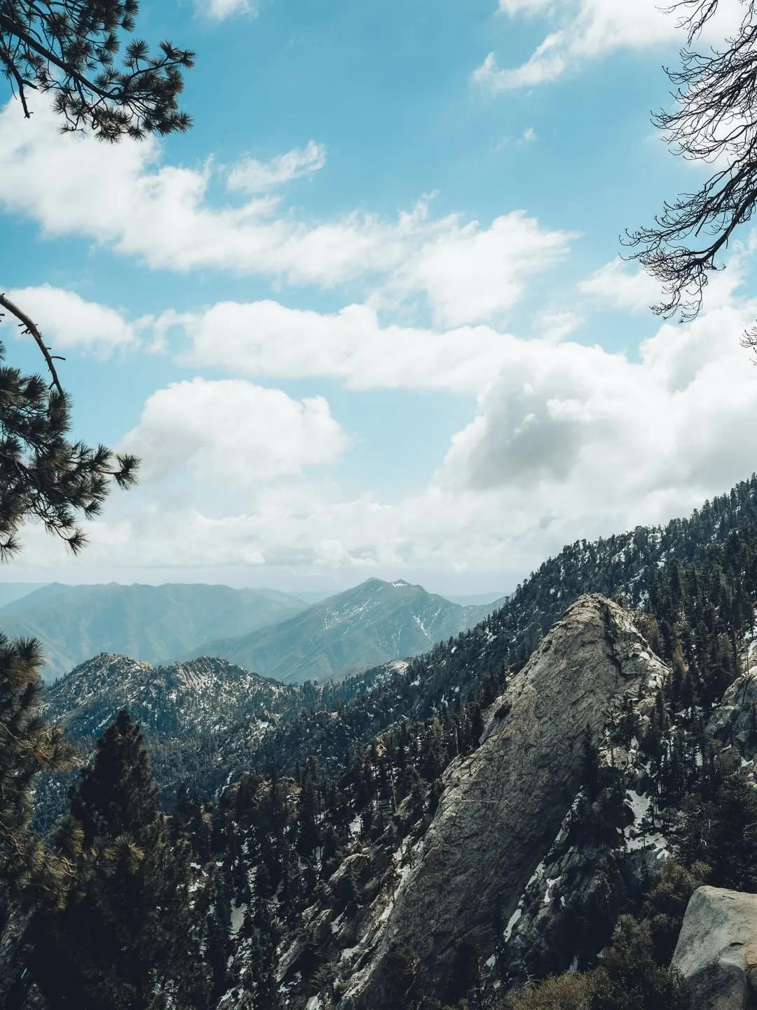 A view across a forested canyon from a high vantage point. A large bare granite outcropping in the foreground on the right, pine trees on the canyon walls, and layered mountain ridges fading into haze under a sky of scattered cumulus.