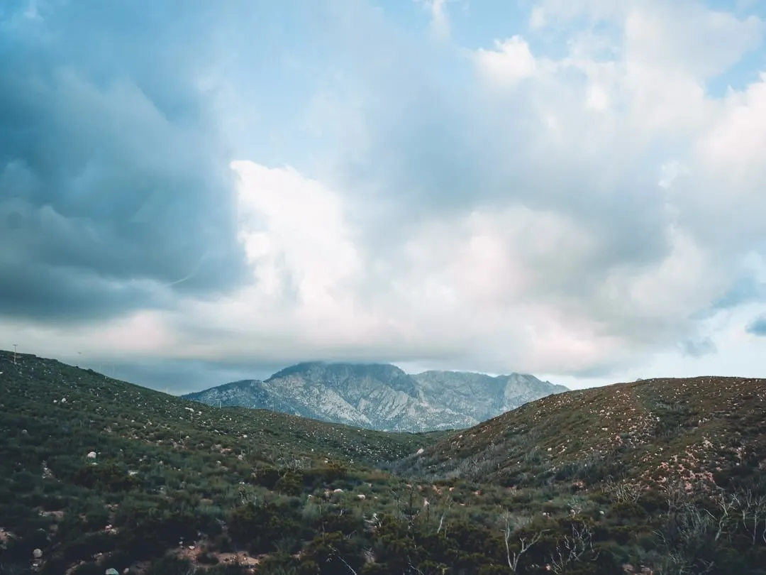 A wide sky of thick cumulus over rolling chaparral hills. A bare granite peak sits in the middle distance, draped in a low cloud bank.