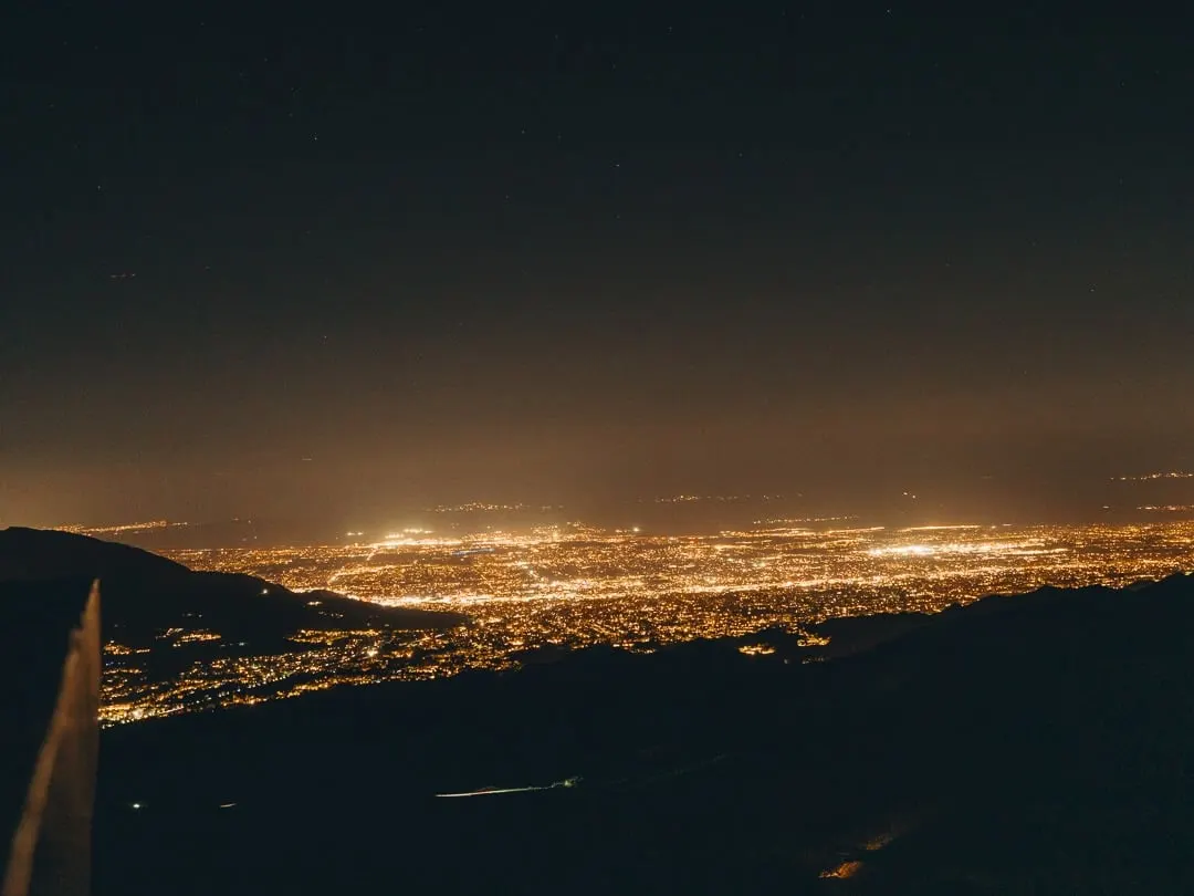 The Coachella Valley at night, seen from a high mountain vantage. An orange grid of city lights spreads to the horizon, dark ridges in the foreground, a faint glow of stars above.