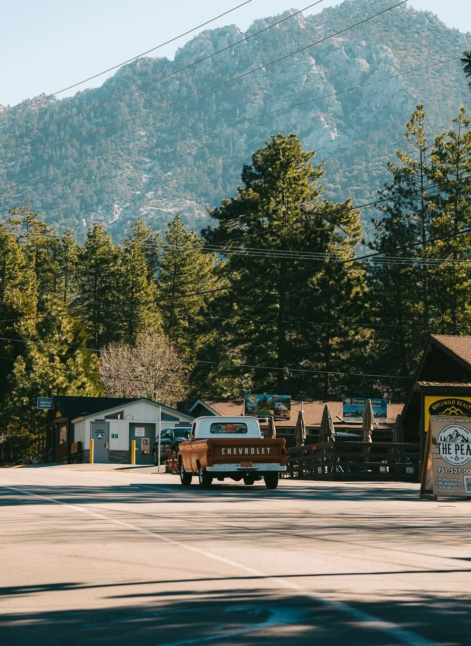 A vintage orange Chevrolet C10 pickup truck driving down a quiet two-lane road through Idyllwild, California. Visitor Center and Idyllwild Realty signs flank the road; pine trees and Tahquitz Peak rise behind.